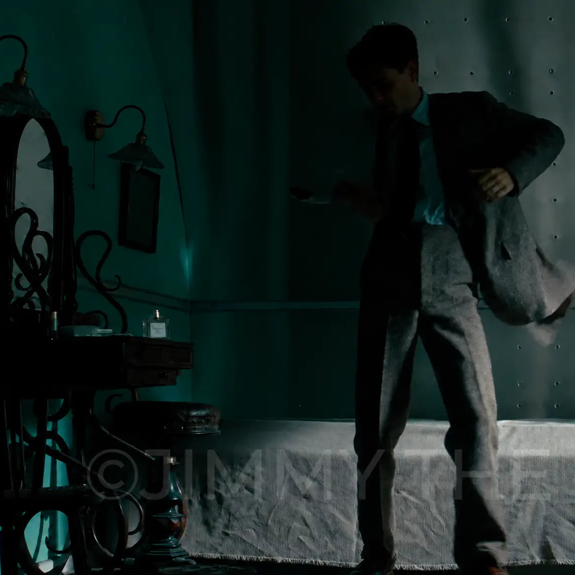 A man in a dark suit and tie stands in a dimly lit room with teal lighting, holding a jacket and looking down, with a mirror, wall sconces, and a bed visible in the background.